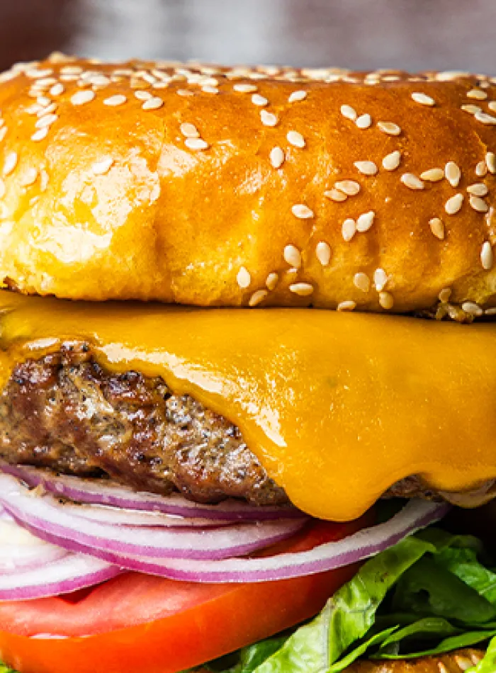 Close-up of a cheeseburger with melted cheddar, red onions, tomato, lettuce, and a sesame seed bun.