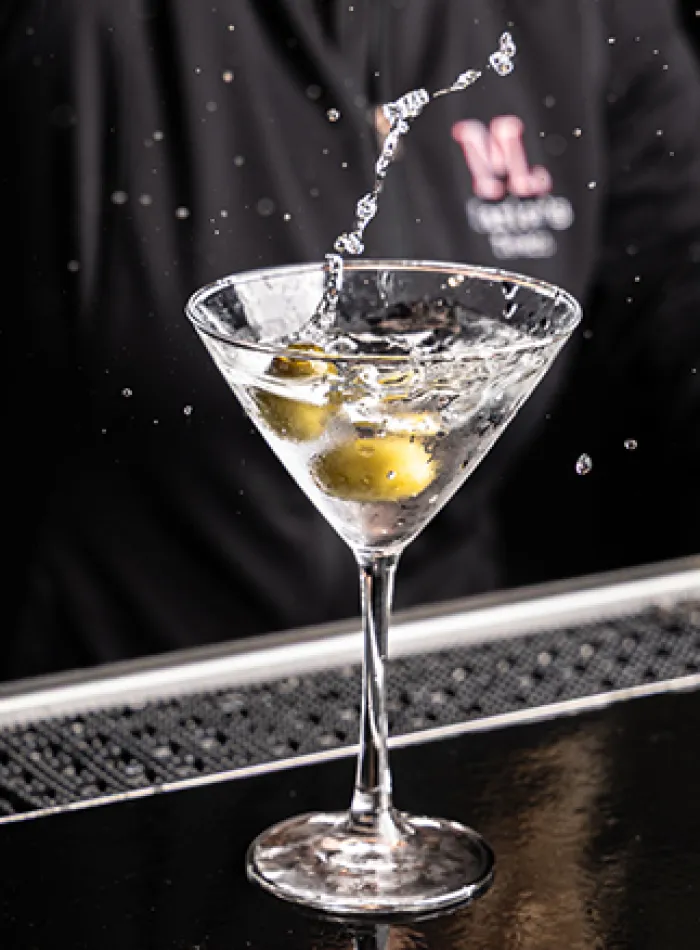 Clear martini glass with olives and water splash on a black bar counter and blurred bartender background.