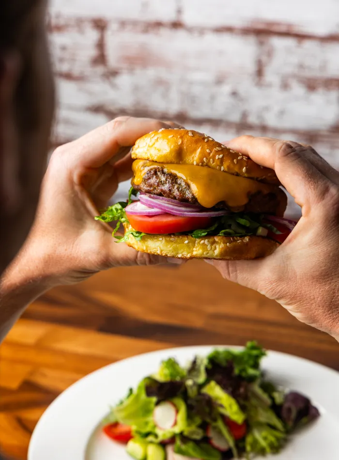 Person holding a cheeseburger with lettuce, tomato, onion, and sesame seed bun over a plate of fresh salad.