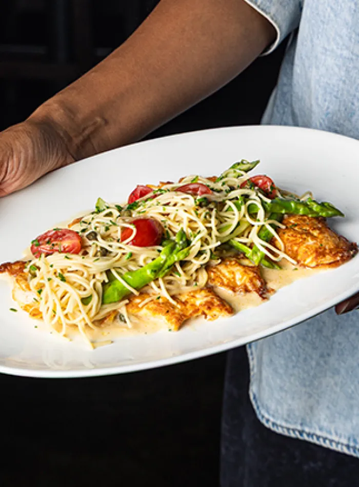 Plate of pasta with cherry tomatoes, asparagus, and grilled chicken served on a white dish held by a person.