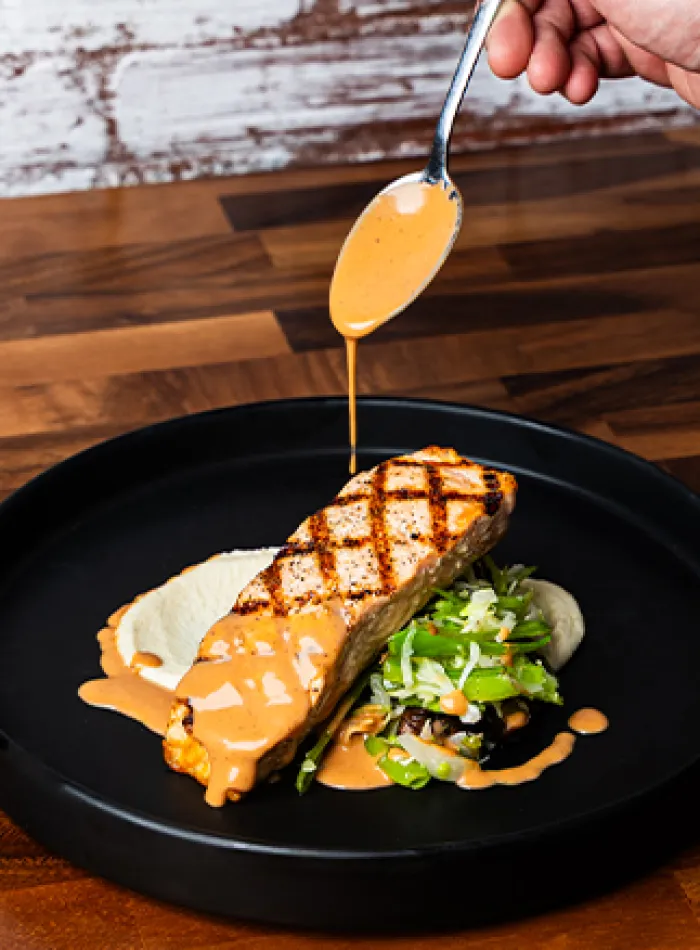 Grilled salmon steak with creamy sauce being poured over it, served with mashed potatoes and sautéed vegetables.