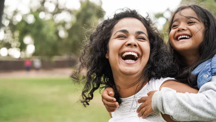 Smiling mother giving piggyback ride to happy daughter outdoors in a park with greenery background