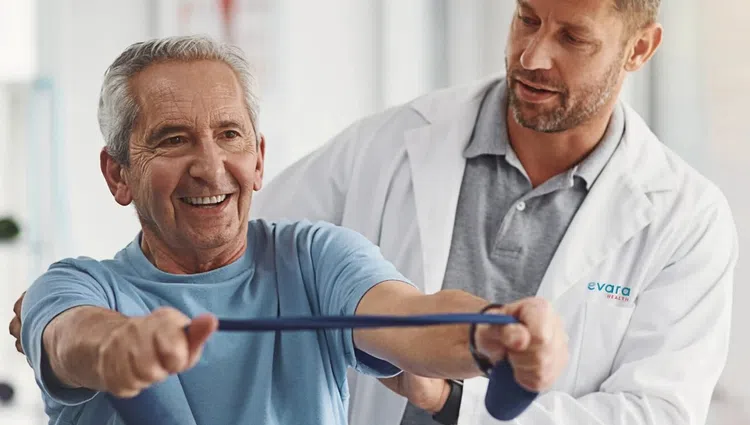 Senior man doing resistance band exercise with a physiotherapist assisting him in a bright clinic.