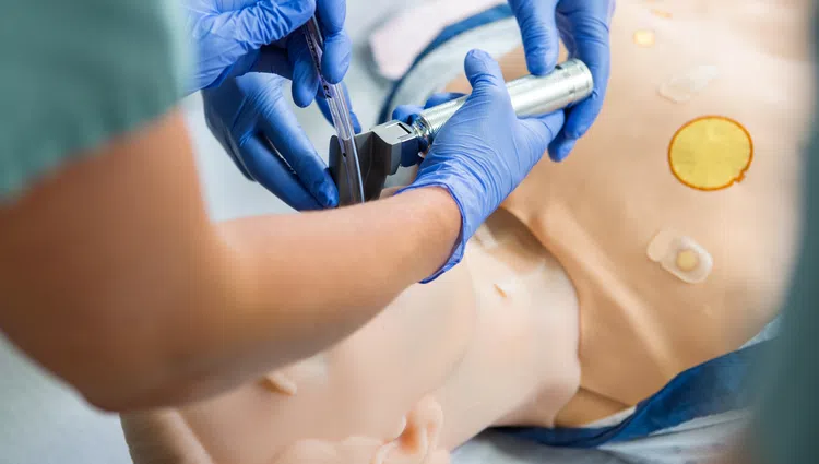 Medical professionals in blue gloves performing a procedure on a training mannequin's airway using a laryngoscope.