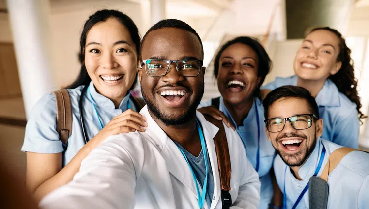 Diverse group of smiling medical students and doctor taking a cheerful selfie indoors, wearing scrubs and lab coats.