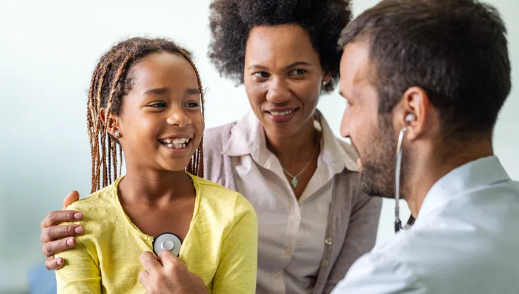 Doctor using stethoscope to check smiling girl's heartbeat with mother supporting her in clinic
