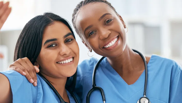 Two diverse female medical professionals smiling, wearing blue scrubs and stethoscopes, looking cheerful and friendly.