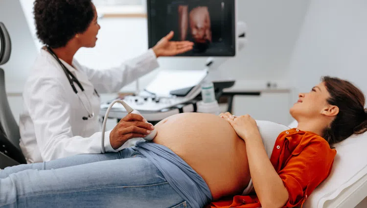 Pregnant woman lying on exam table while doctor performs ultrasound and shows 3D baby image on screen