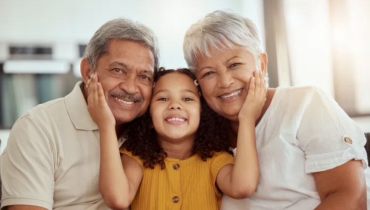 Happy granddaughter smiling and hugging her elderly grandparents in a warm, bright living room setting.