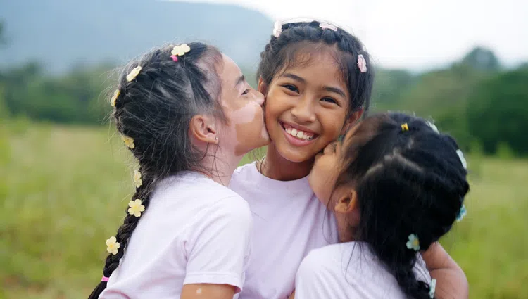 Three young girls smiling and hugging outdoors with flowers in their braided hair and a natural background.