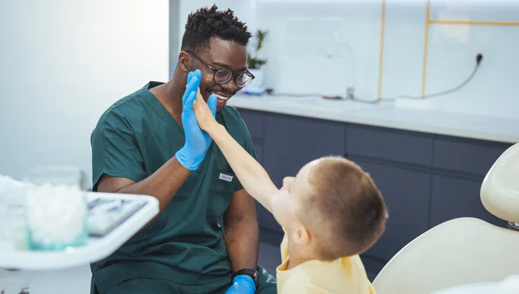 Smiling dentist in green scrubs gives a high five to a young boy in a dental clinic.