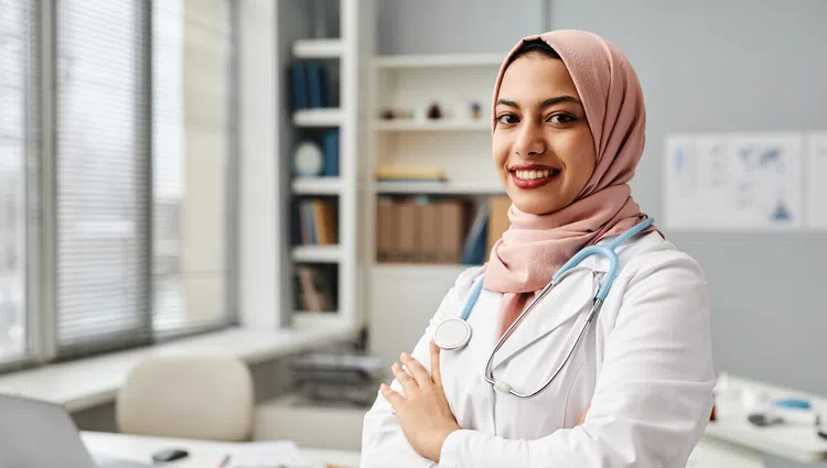 Smiling female doctor in pink hijab and white coat with stethoscope in modern medical office with laptop and shelves.