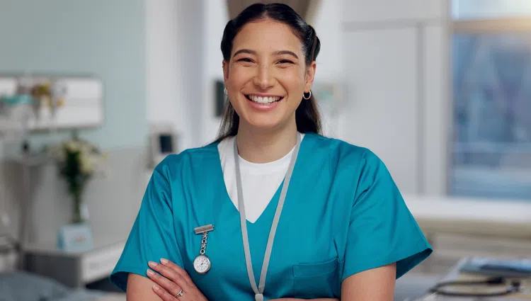 Smiling female nurse in blue scrubs standing confidently in a bright medical clinic room with arms crossed.