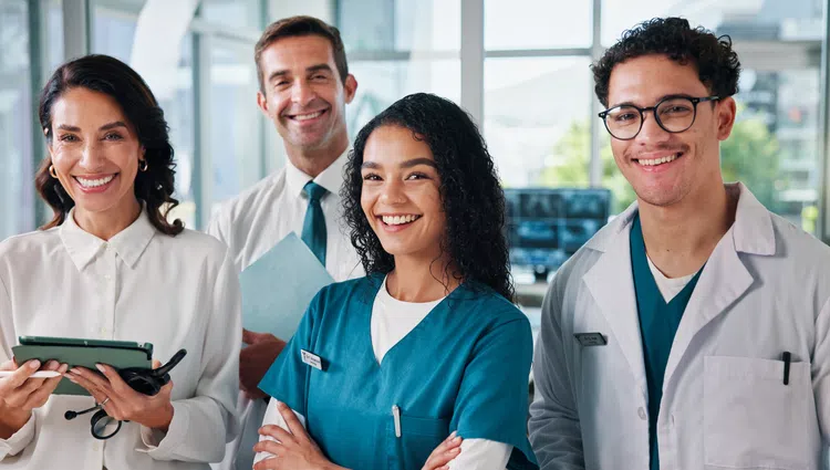 Smiling diverse healthcare professionals posing confident in hospital environment with tablets and medical uniforms.
