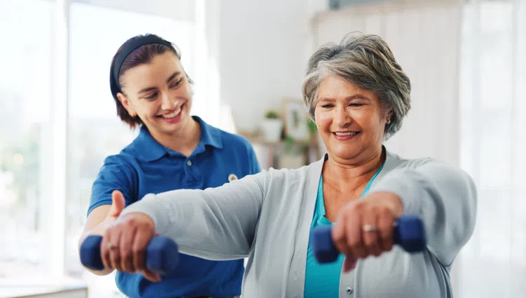 Senior woman lifting dumbbells with a smiling female trainer assisting her in a bright indoor setting
