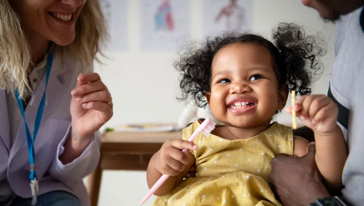 Smiling toddler in yellow dress holding toothbrushes with adults showing dental care in background