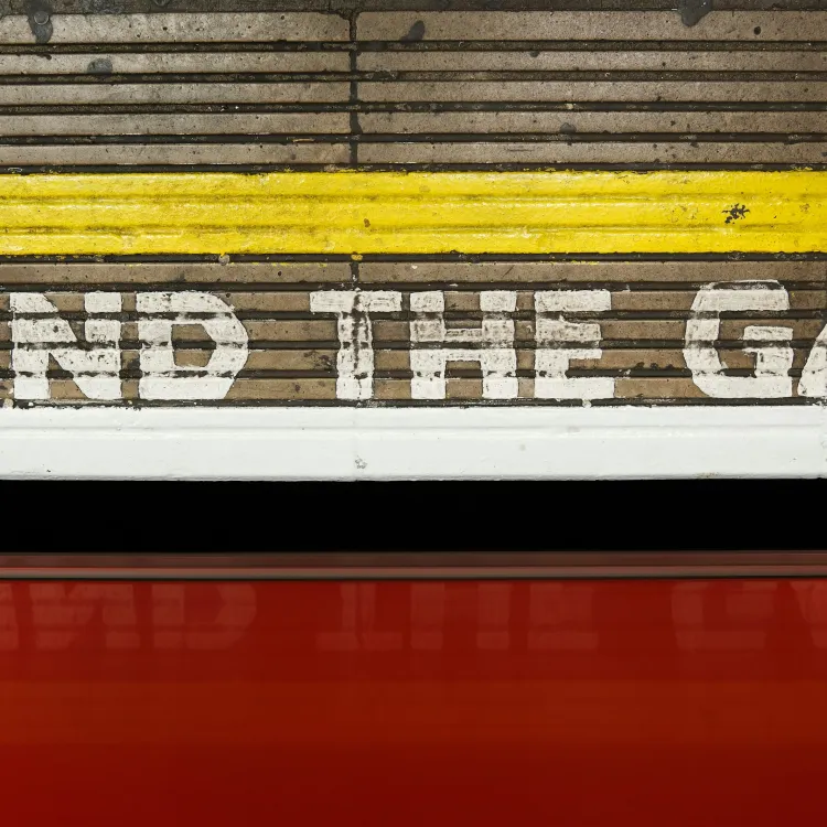 MIND THE GAP painted on wooden train platform edge with red train below and a yellow safety line above.