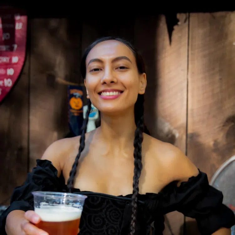 Smiling woman with braided hair holding a beer in a plastic cup against a rustic wooden background.
