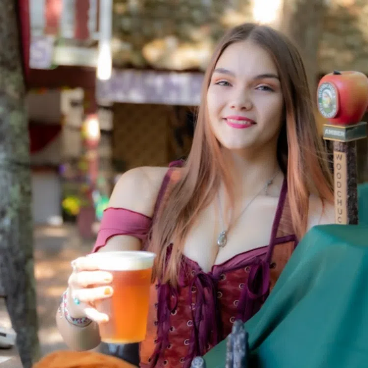 Smiling young woman holding a glass of beer at an outdoor festival or market during daytime.