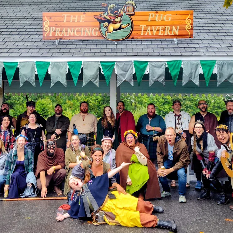 Group of people in colorful medieval costumes posing outside The Prancing Pug Tavern with green bunting overhead.