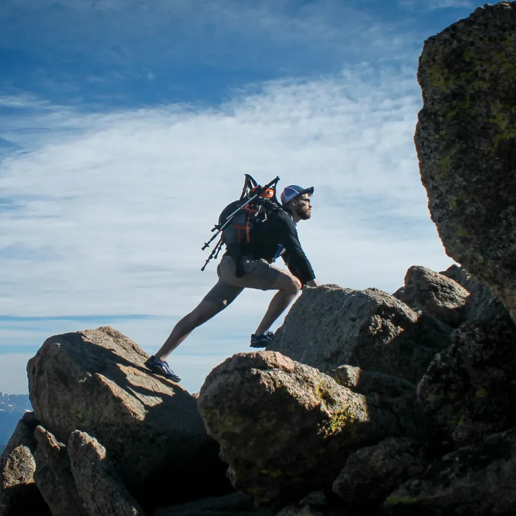 Hiker with backpack and trekking poles climbing rocky mountain under blue sky with distant snowy peaks