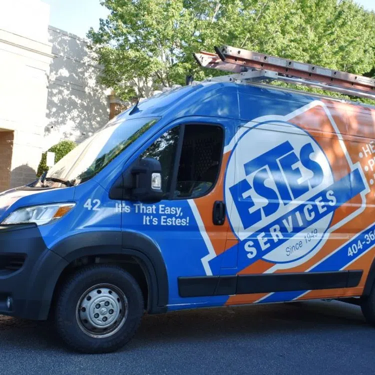 Estes Services blue and orange service van parked outside Estes Heating & Air Conditioning building with technician standing beside it