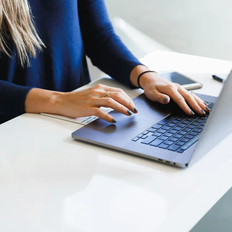 Person typing on a laptop keyboard at a white desk with a smartphone nearby in natural light.