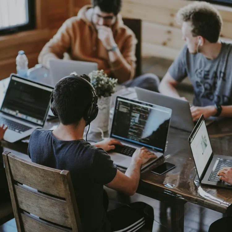 Group of young people working on laptops around a wooden table in a casual office setting.