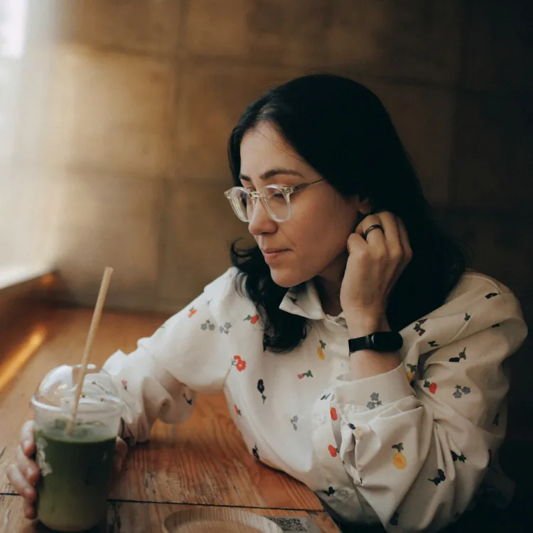 Woman in glasses and floral shirt sitting by a window holding a green smoothie in a cozy cafe.