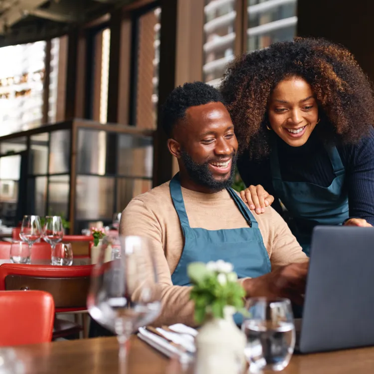 Smiling male and female restaurant workers in aprons using a laptop in a modern dining setting.