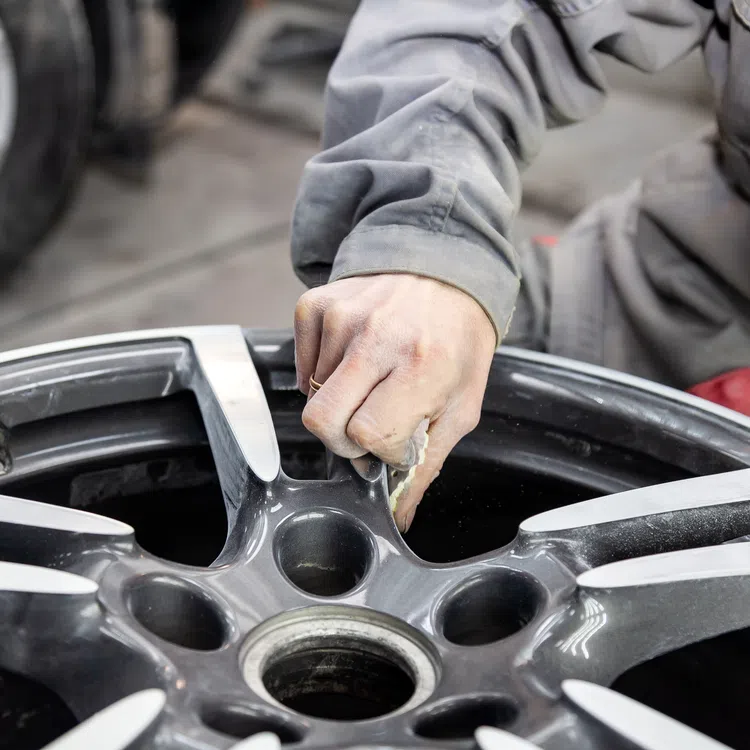 Mechanic cleaning a black alloy car wheel rim with a cloth in an auto repair shop.