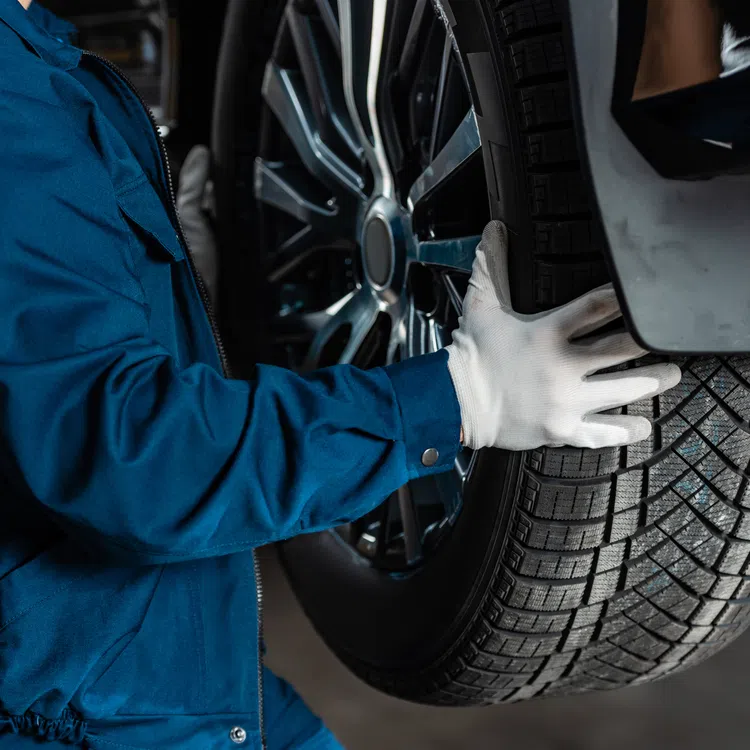 Mechanic in blue uniform installing or removing a car tire with gloved hands in an auto repair shop.