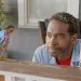 A man looks thoughtfully at a small animated character beside him on a porch.
