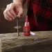 Hand applying red threadlocker adhesive to a metal bolt standing upright on a wooden surface with a plaid shirt background.