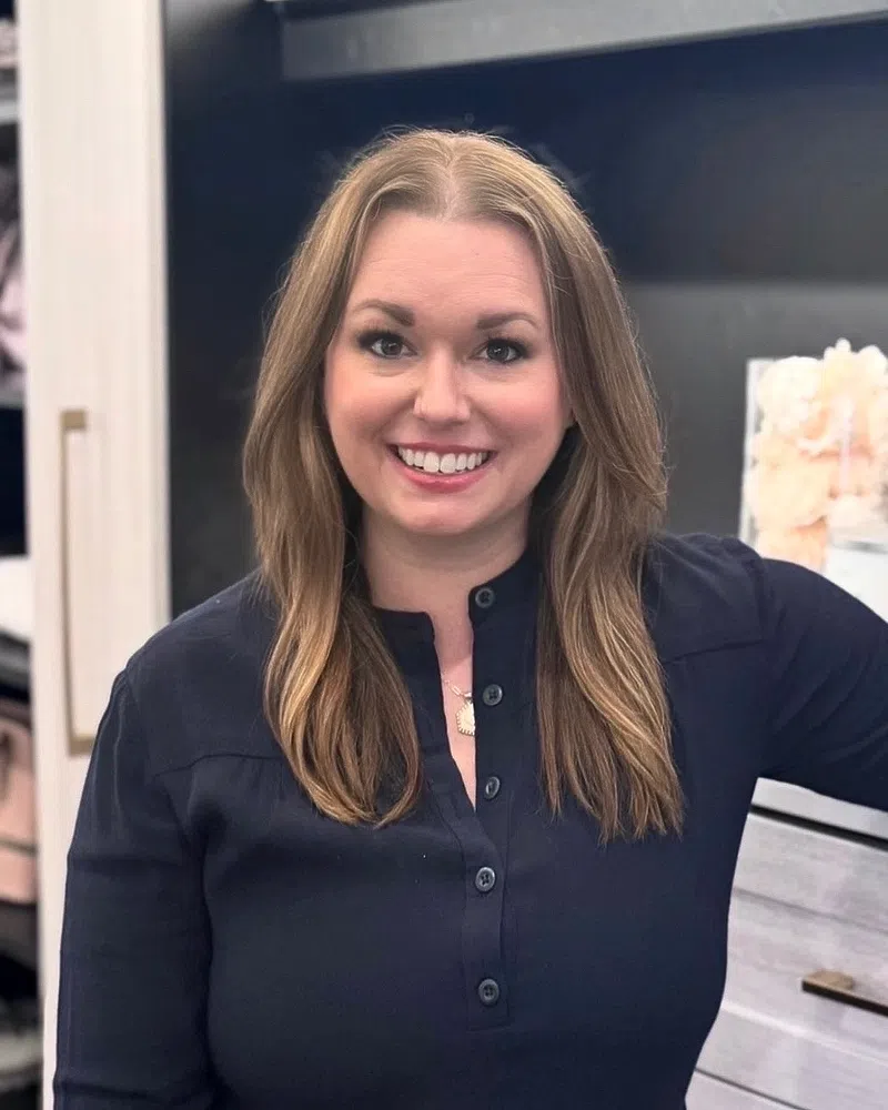 Smiling woman with long light brown hair wearing a dark blouse standing in a modern kitchen with white cabinets.