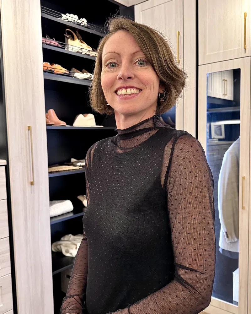 Smiling woman in sheer black polka dot top posing in a modern walk-in closet with shoes and clothes.