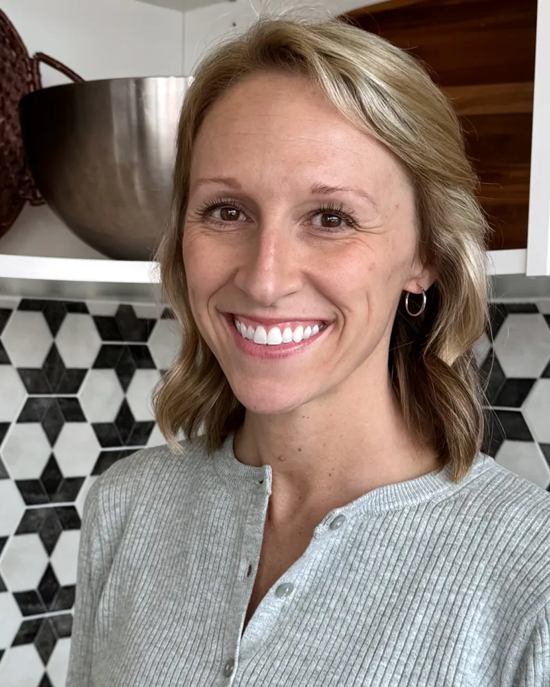 Smiling woman with blonde hair wearing a gray sweater in a kitchen with black and white tile backsplash.