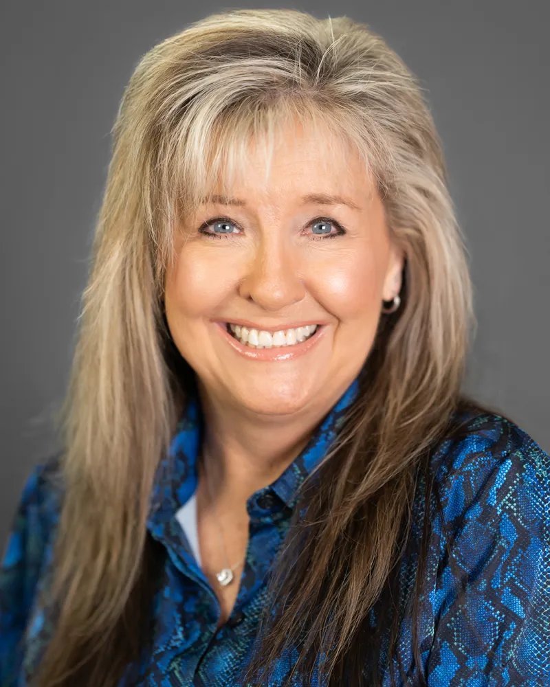 Smiling middle-aged woman with long blonde hair wearing a blue patterned blouse against a gray background