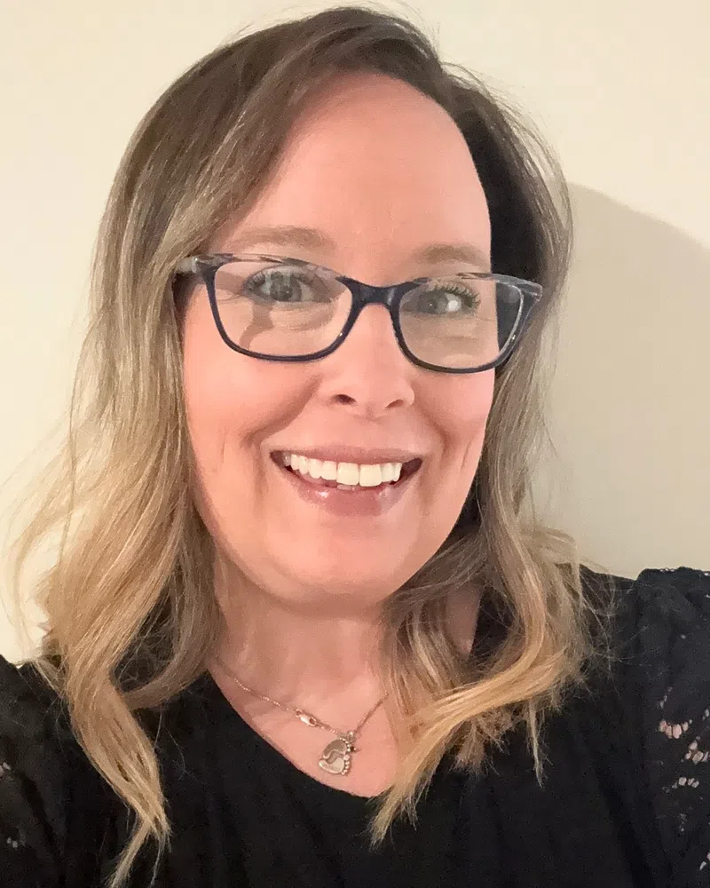 Smiling woman with glasses and wavy blonde hair wearing a black lace top and heart necklace indoors.