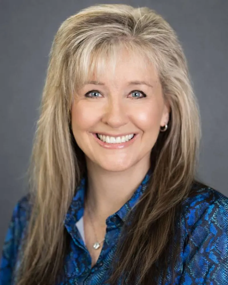 Smiling woman with long blonde hair wearing a blue patterned blouse against a plain gray background