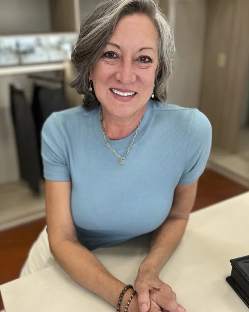 Smiling mature woman with gray hair wearing a blue shirt leaning on a table in a cozy room.