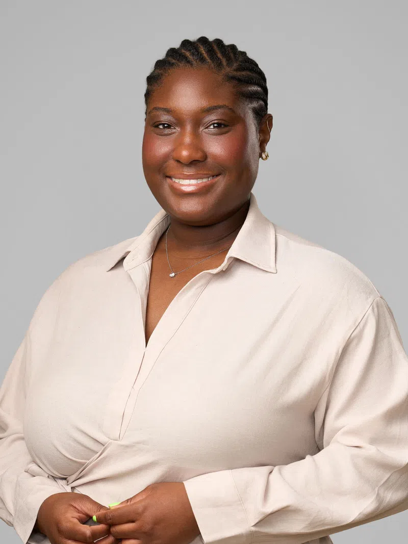 Smiling Black woman with braided hair wearing a beige shirt and small necklace against a plain gray background