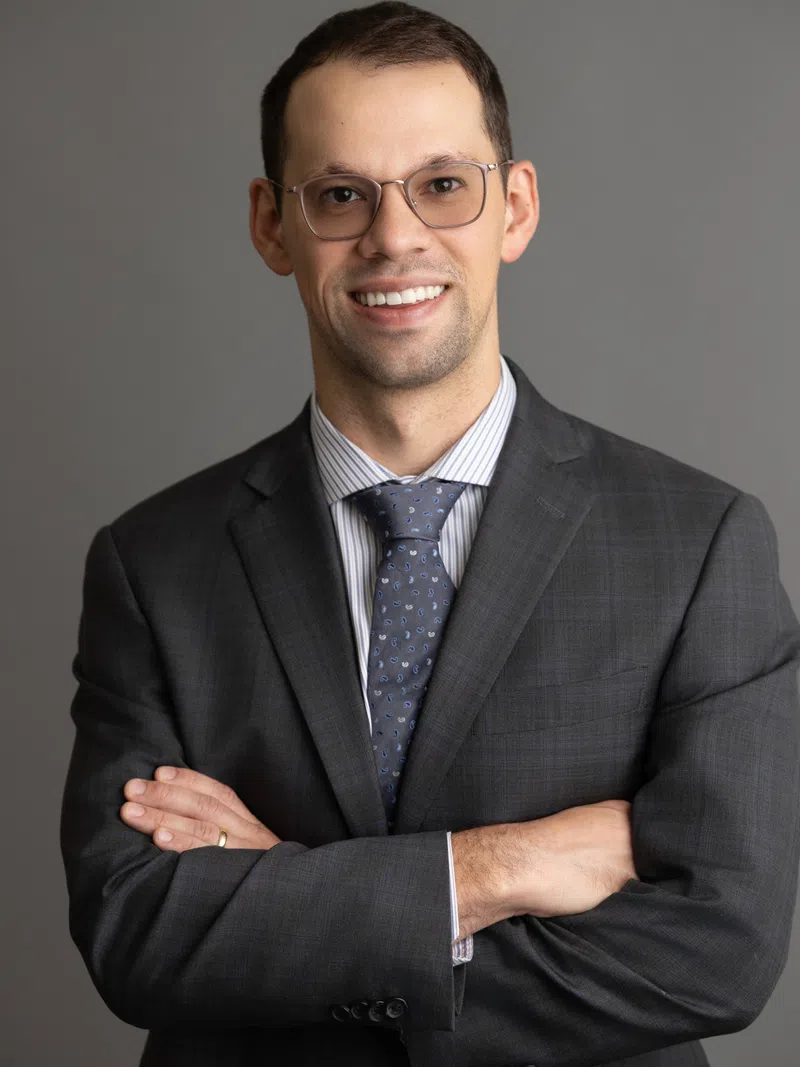 Smiling man in glasses wearing a gray suit and patterned tie standing with arms crossed against gray background