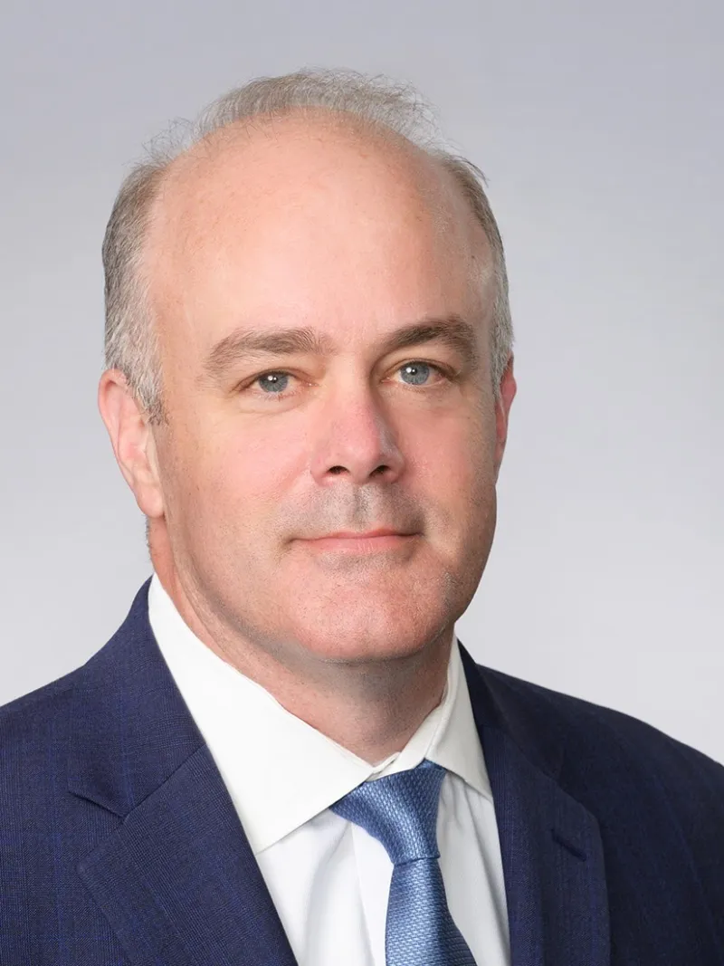 Professional headshot of a man in a navy suit and light blue tie against a gray background.
