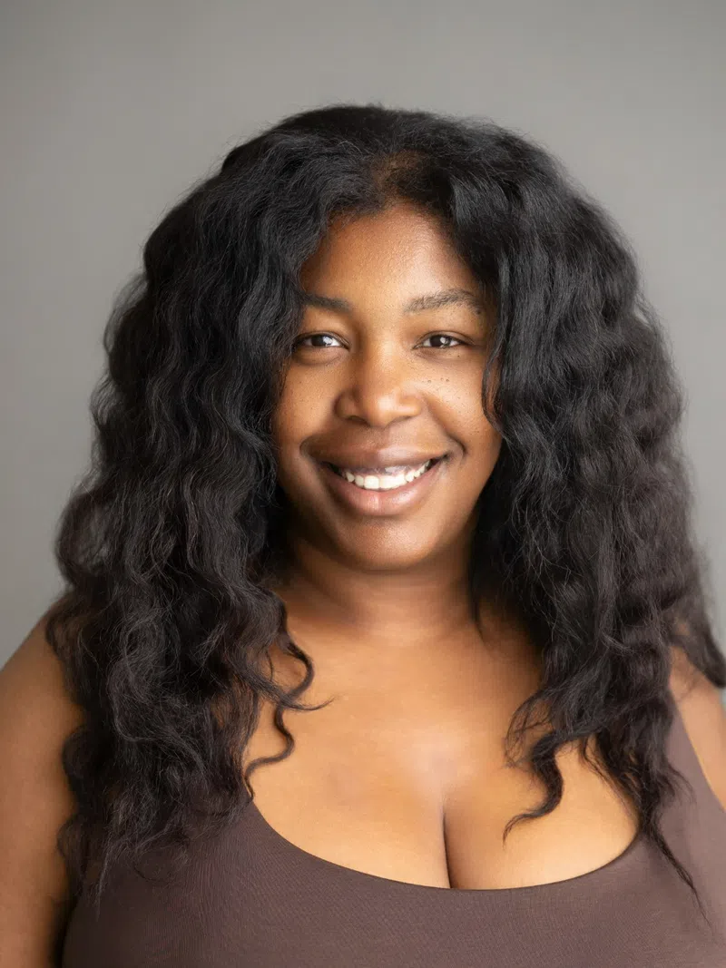 Smiling woman with long curly black hair wearing a brown sleeveless top on gray background.