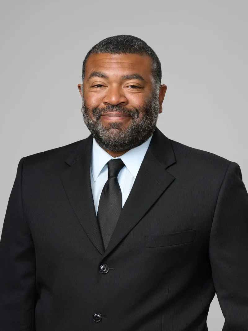 Confident man in black suit and tie smiling against a plain gray background, professional studio portrait.