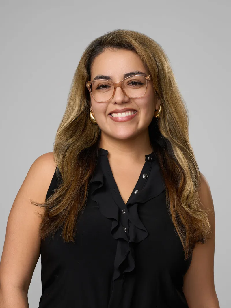 Smiling woman with long highlighted hair wearing glasses and a black ruffled sleeveless top against gray background