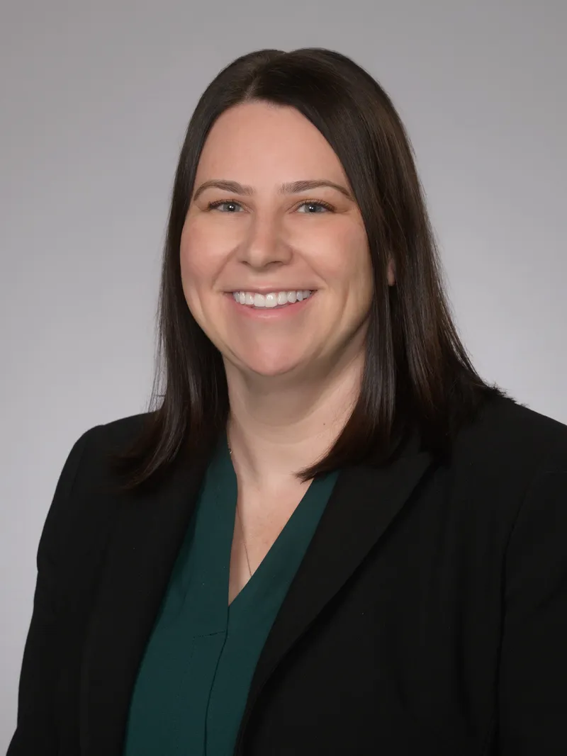 Smiling professional woman with straight dark hair wearing a black blazer and green blouse on gray background