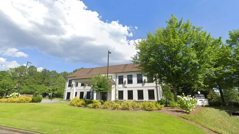Two-story white office building with brown roof surrounded by green trees and manicured lawn under blue sky