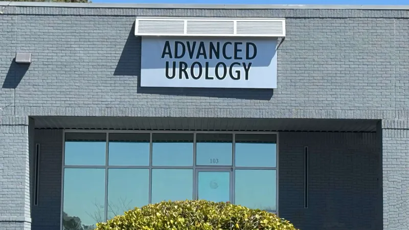 Exterior of Advanced Urology clinic with large front windows and a trimmed bush under clear blue sky.
